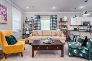 Una sala de estar con sofá, sillones, una mesa de centro y una zona de cocina con armarios y electrodomésticos.