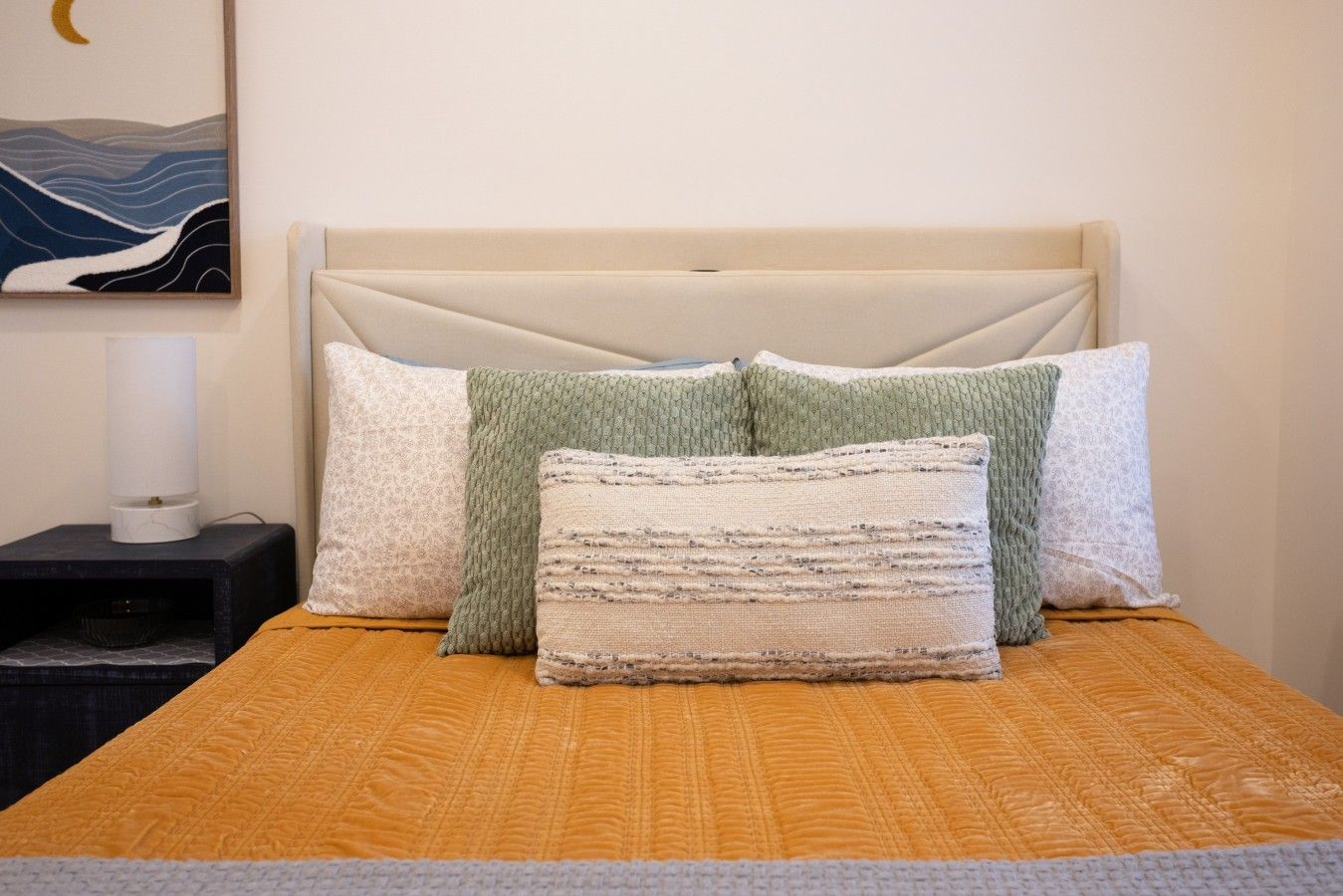 Una cama con cabecera color crema, colcha naranja con textura y tres almohadas en tonos verdes y blancos.