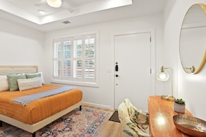 Un dormitorio con cama, ventana con persianas, puerta, mesita de noche con lámpara y espejo en la pared.