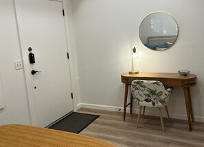 Una habitación con una puerta blanca, un escritorio de madera con una silla, un espejo redondo, una lámpara de pie y una mesita con un cuenco.