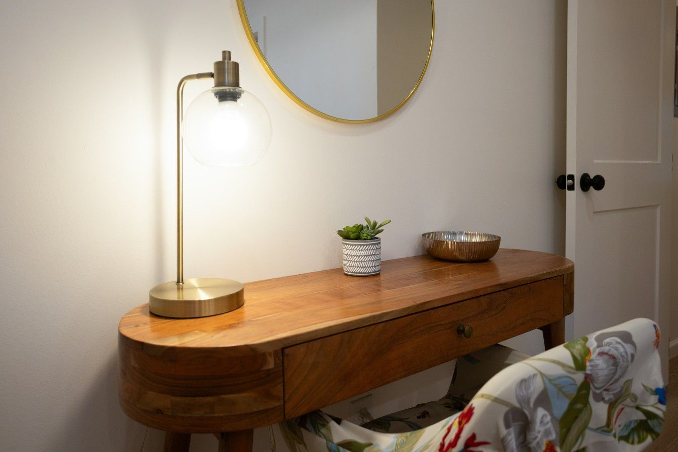 Une coiffeuse en bois avec un miroir rond, une lampe sur pied, une petite plante en pot et un bol décoratif.