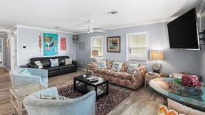A living room with a black sofa, a floral sofa, a glass coffee table, and a TV mounted on the wall. - Snowbird-Friendly 3BR Gulf Shores Home Near Beach (Gulf Shores)