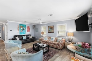 Ein Wohnzimmer mit einem schwarzen Sofa, einem Blumensofa, einem Glastisch und einem an der Wand befestigten Fernseher.