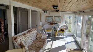 Una habitación soleada con un sofá floral, sillas de mimbre, una mesa con un florero y vistas a través de puertas de cristal.