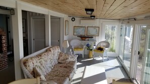Ein sonnendurchfluteter Raum mit einem blumigen Sofa, Korbstühlen, einem Tisch mit einer Vase und Blick durch Glastüren.