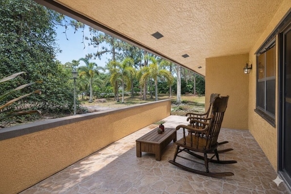 A covered patio with a rocking chair, a small table, and a view of tropical trees.