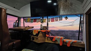 Interno di un veicolo con un'ampia finestra, un televisore montato a soffitto e vista su un paesaggio al tramonto.