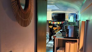 Un corridoio stretto con pavimento in legno, una porta con specchio circolare e una piccola zona relax con una sedia e un tavolino.
