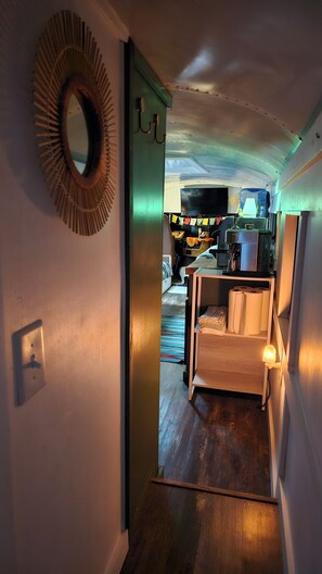 Un estrecho pasillo con suelo de madera, una puerta con un espejo circular y una pequeña zona de estar con una silla y una mesa.