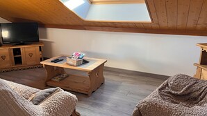 Een loft-achtige kamer met een schuin dak, een dakraam, een televisie op een houten kast, een klein houten tafeltje met tijdschriften en twee comfortabele stoelen.