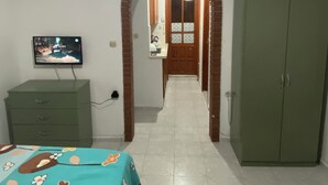 A bedroom with a TV, a wardrobe, a bed with a floral bedspread, and a hallway leading to a bathroom. - Beluna City Apart (Marmara)