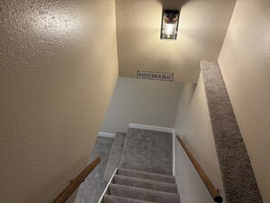 A carpeted staircase with a handrail, a wall-mounted light fixture, and a warning sign. - 2 King Suites & A Secret Bookcase Room + Pool Table & Ping Pong, AC (Coeur d'Alene)