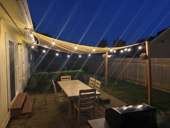 Une terrasse extérieure avec une table et des chaises, des guirlandes lumineuses et un barbecue.