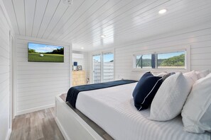 Un dormitorio con cama, televisor montado en la pared, ventana con persianas y vistas al exterior.