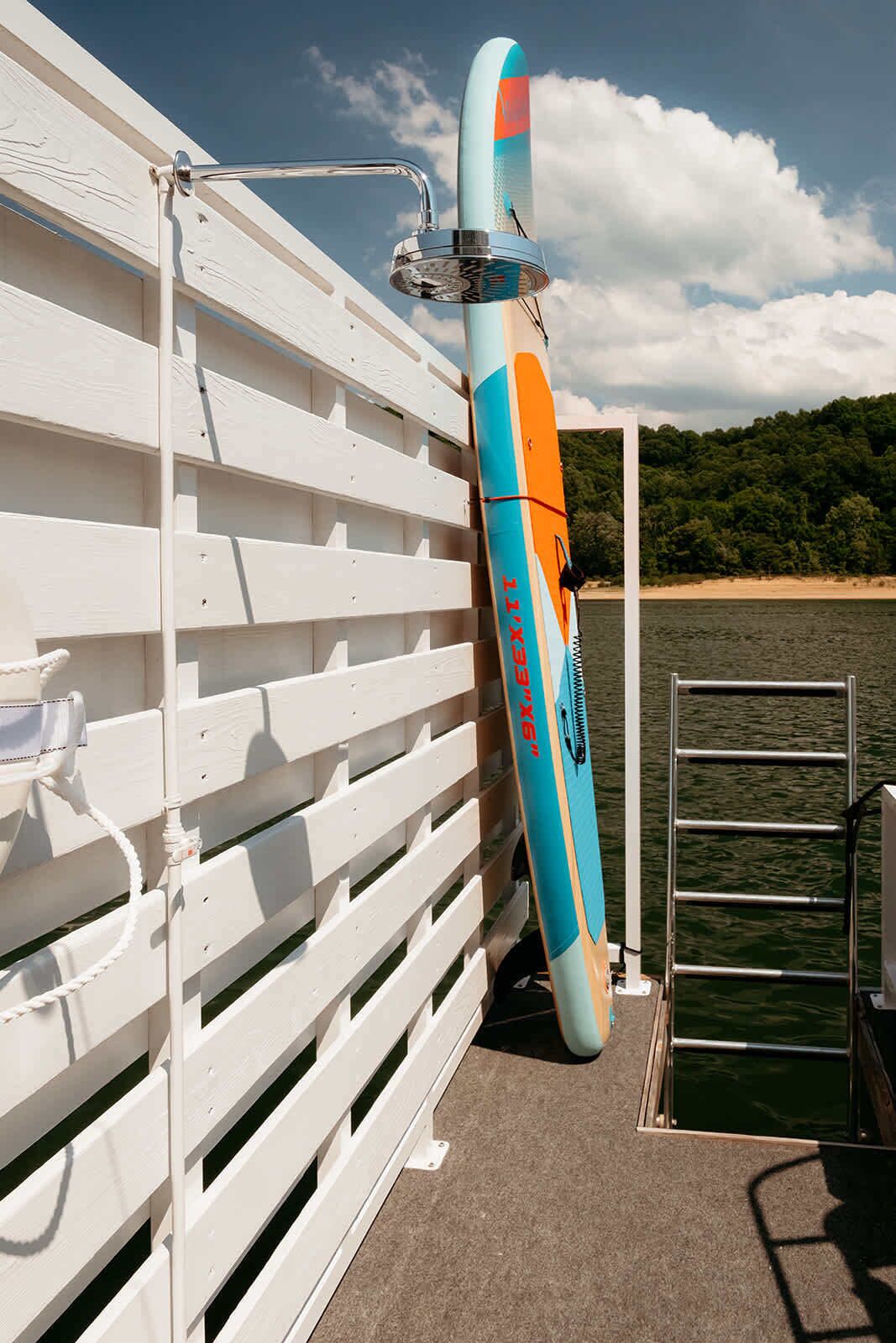 Een witte houten omheining, een blauw en oranje surfplank ertegen, en een metalen ladder die naar een steiger leidt.