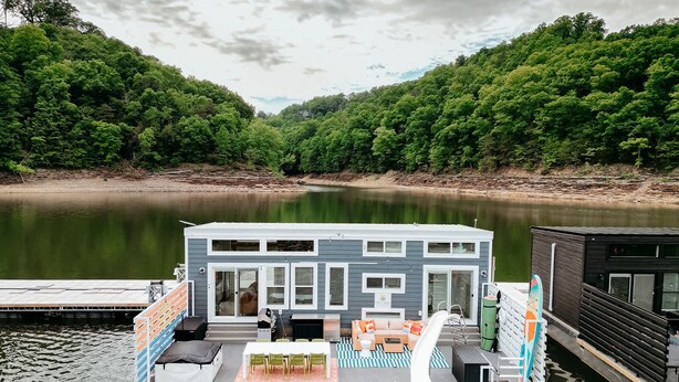 Une maison flottante dotée d’une terrasse, de coins salon extérieurs et offrant une vue sur une rivière paisible et une forêt.