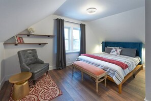 A bedroom with a bed, a chair, a bench, a small table, and a window with curtains. - HNU  · HNU  NEWLY Decorated 2 Bedroom + Den Apartment (Kitchener)