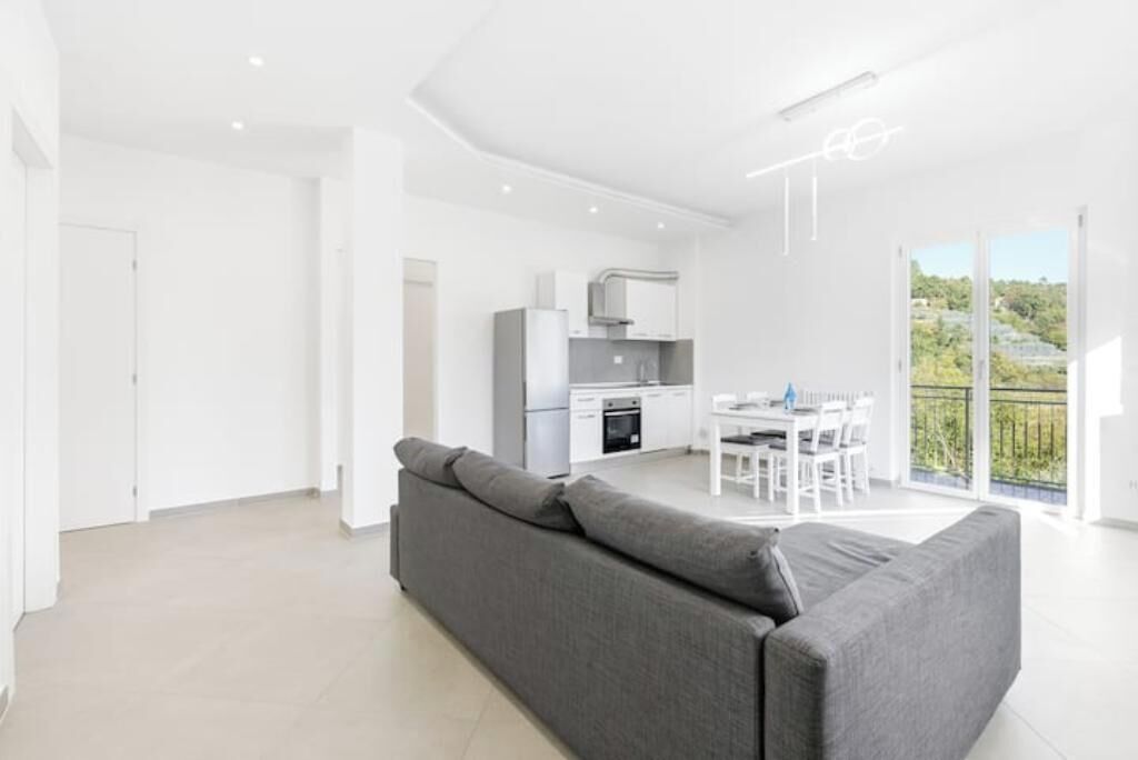 Ein moderner Wohnbereich mit grauem Sofa, einer Küche mit weißen Geräten und einem Essbereich.