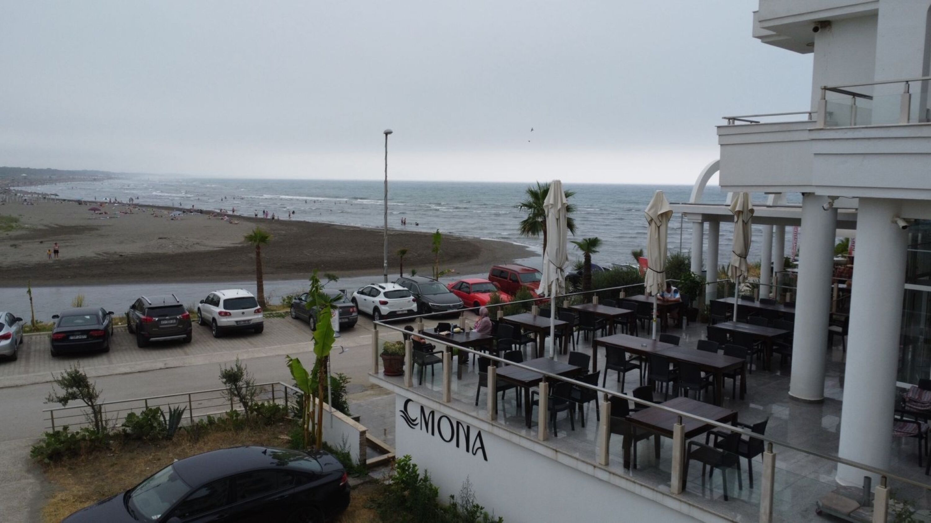 A seaside view with a beach, a parking area, and a restaurant named 'CMONA' with outdoor seating.