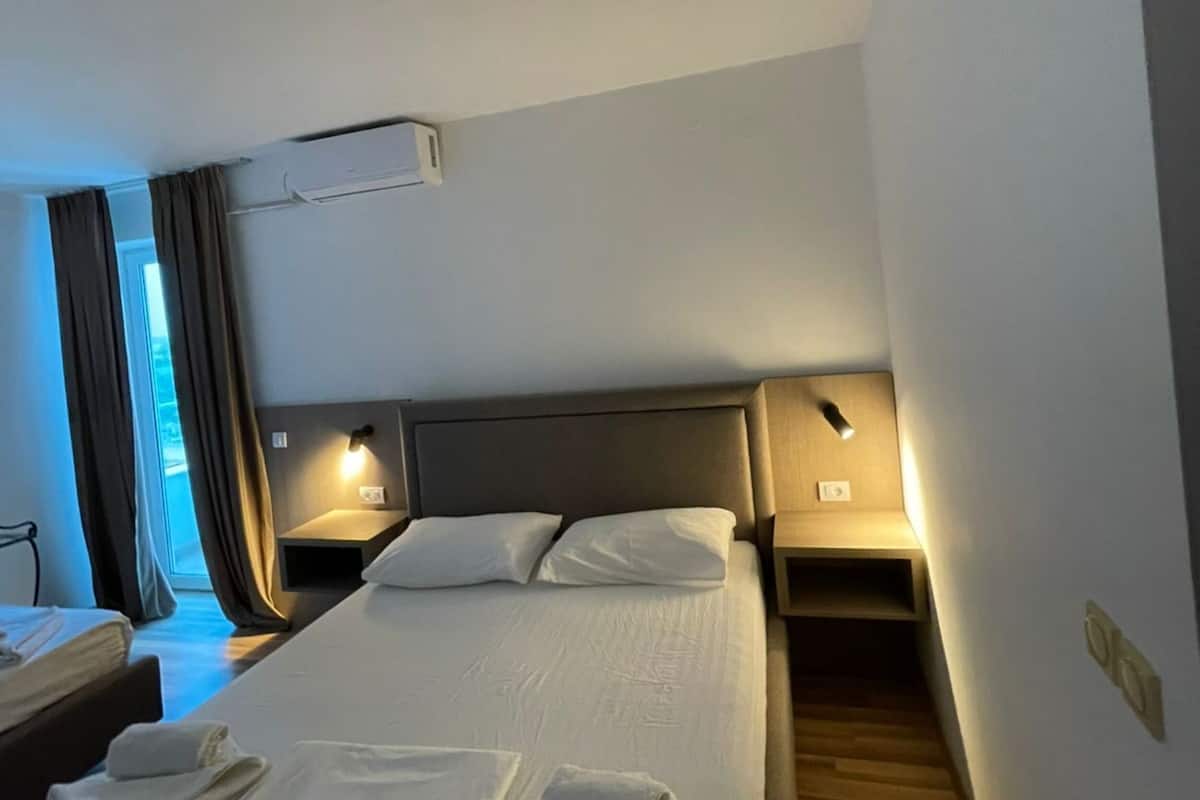 A hotel room with a large bed, bedside tables with lamps, an air conditioning unit, and a window with curtains.