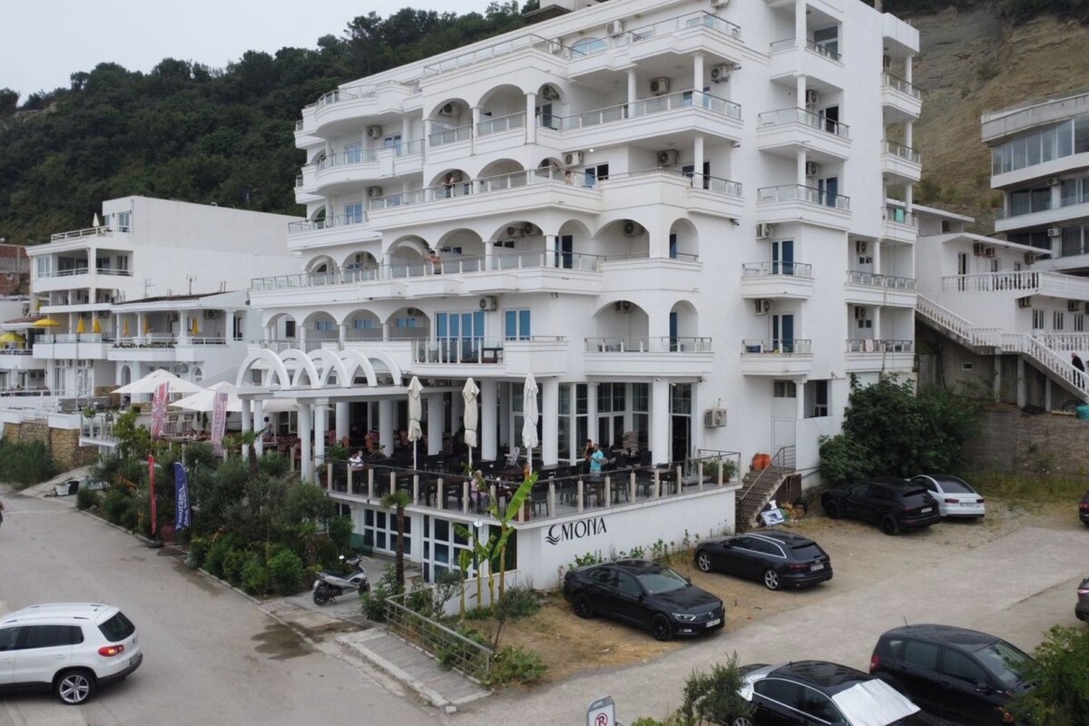A multi-story building with balconies, a restaurant area, and a parking lot with cars.