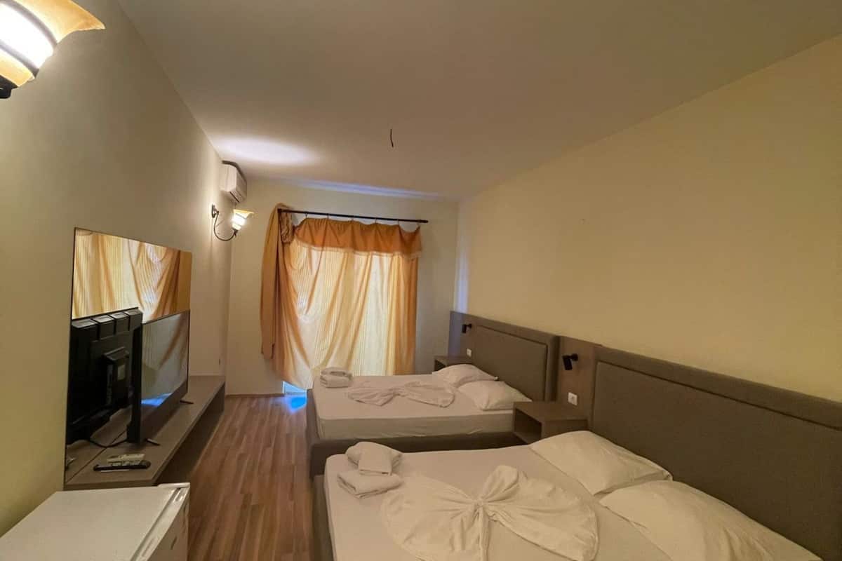 A hotel room with two beds, a TV, a small refrigerator, and a window with curtains.