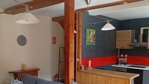 Una sala de estar con sofá, mesa de comedor y sillas, una cocina con baldosas rojas y negras, vigas de madera e iluminación colgante.