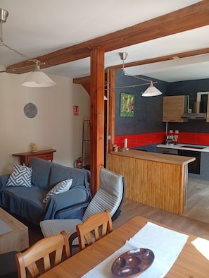 Ein Wohnzimmer mit einer Couch, einem Esstisch und Stühlen, eine Küche mit roten und schwarzen Fliesen, Holzbalken und Pendelleuchten.