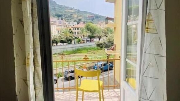 Un balcón con una silla amarilla, un ventanal de vidrio y vista a una zona residencial con edificios y automóviles.