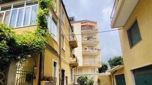 Una calle estrecha bordeada por edificios de varios pisos con balcones, una reja y algo de vegetación.