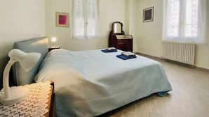 Un dormitorio con cama, lámpara de noche, cómoda, espejo, ventanas y un radiador.