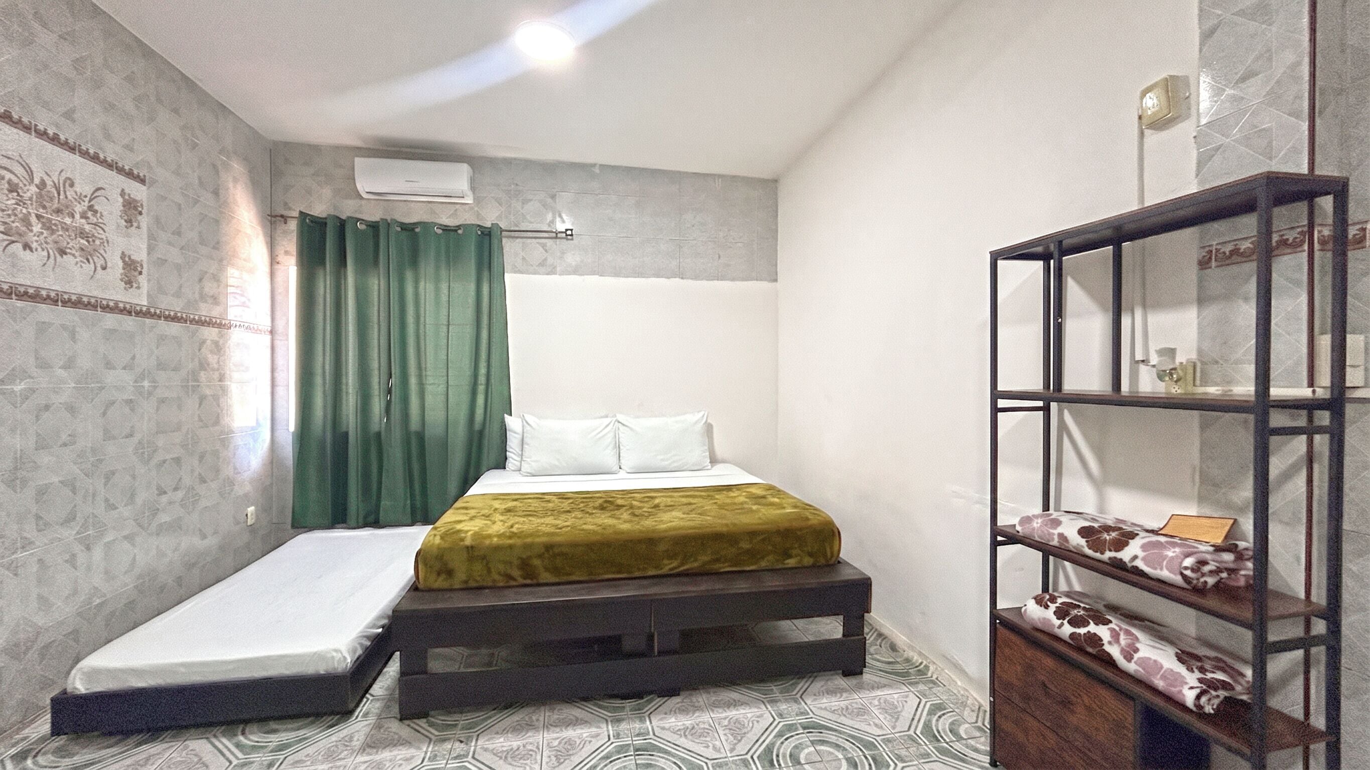 Quarto de hotel compacto com cama, pequena mesa de cabeceira, guarda-roupa e ar condicionado fixado na parede.