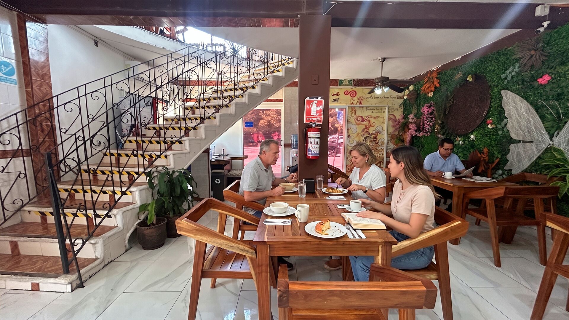 A restaurant with wooden tables and chairs, a staircase with metal railings, and a decorative wall with plants and a butterfly.