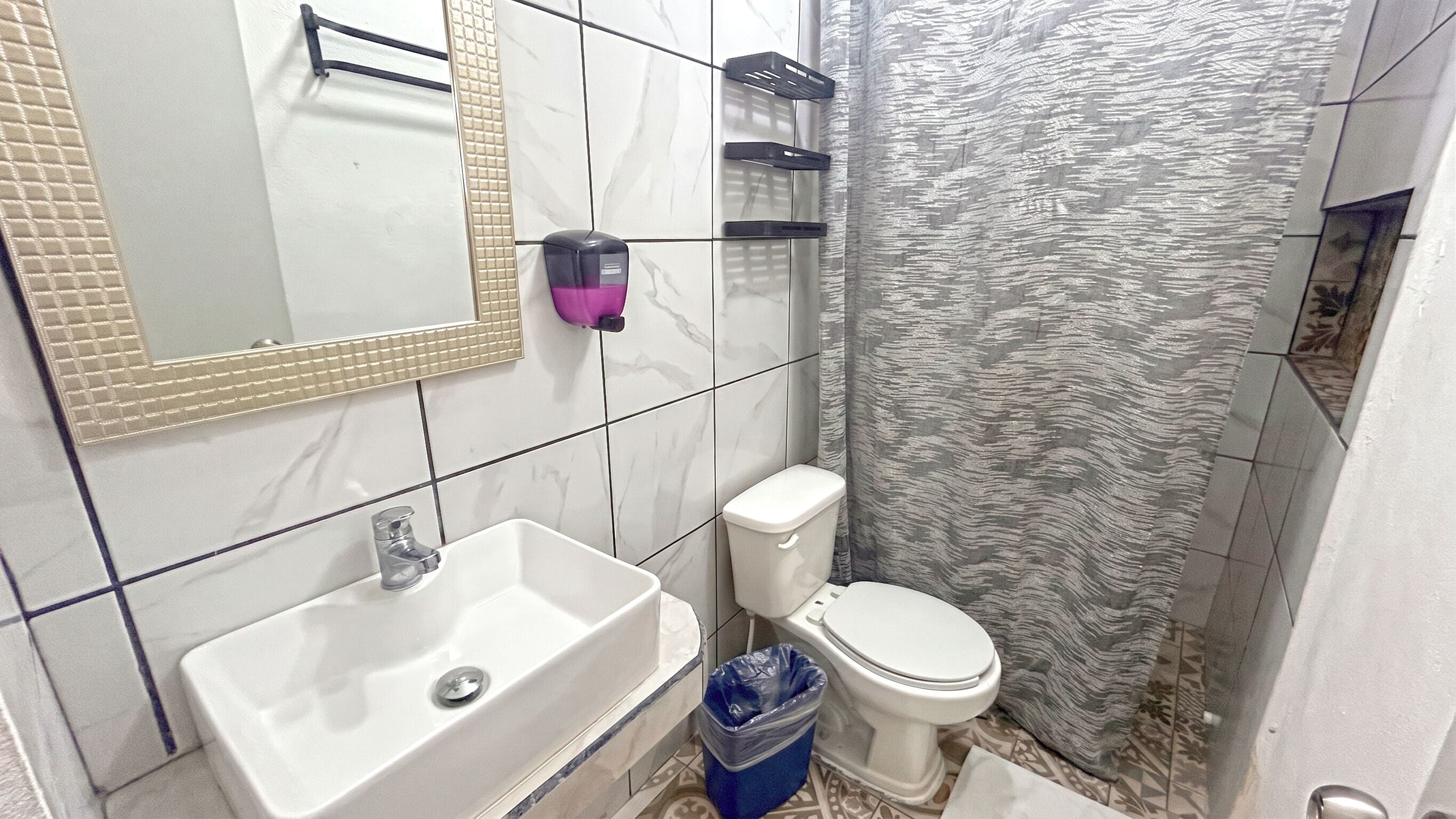 Ein Badezimmer mit einem weißen Waschbecken, einer Toilette, einem Spiegel mit dekorativem Rahmen und einer Duschvorhang mit strukturierter Musterung.