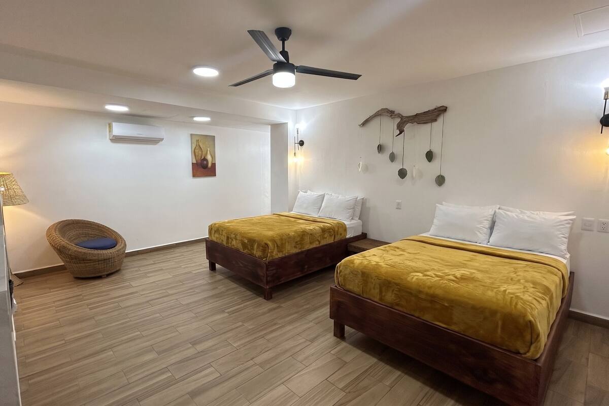 Habitación de hotel con dos camas, ventilador de techo, iluminación empotrada, aire acondicionado de pared y una silla de mimbre.