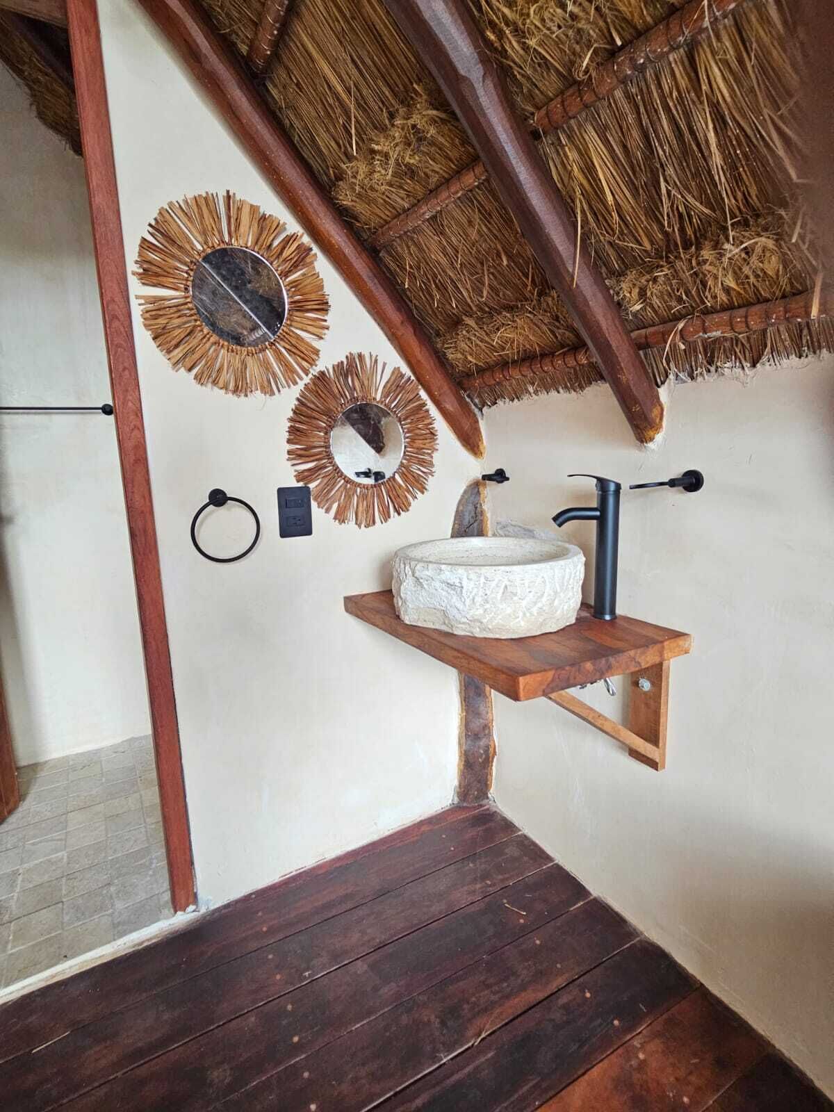 A rustic bathroom with a wooden shelf holding a stone basin, a metal faucet, and two wall-mounted mirrors with straw-like frames.