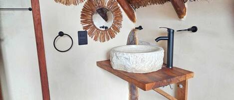A rustic bathroom with a wooden shelf holding a stone basin, a metal faucet, and two wall-mounted mirrors with straw-like frames.