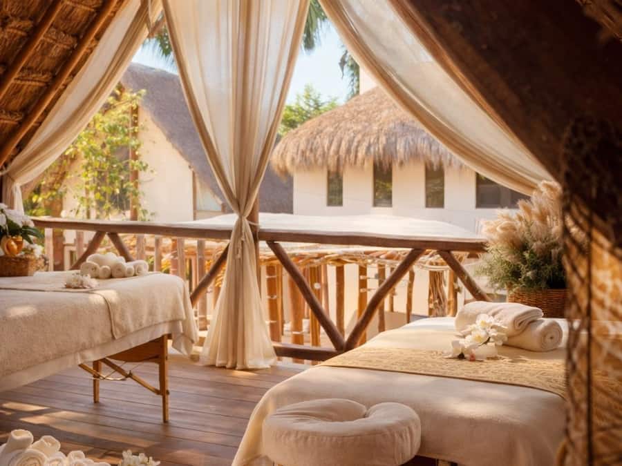 A cozy, well-lit room with a wooden ceiling, a bed with a white bedspread, a small wooden bench, and a wicker basket with towels and a candle.