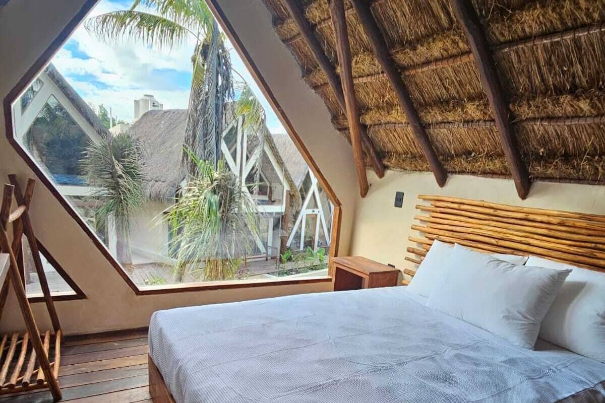 Un dormitorio con cama, piso de madera, techo de paja y un ventanal grande.