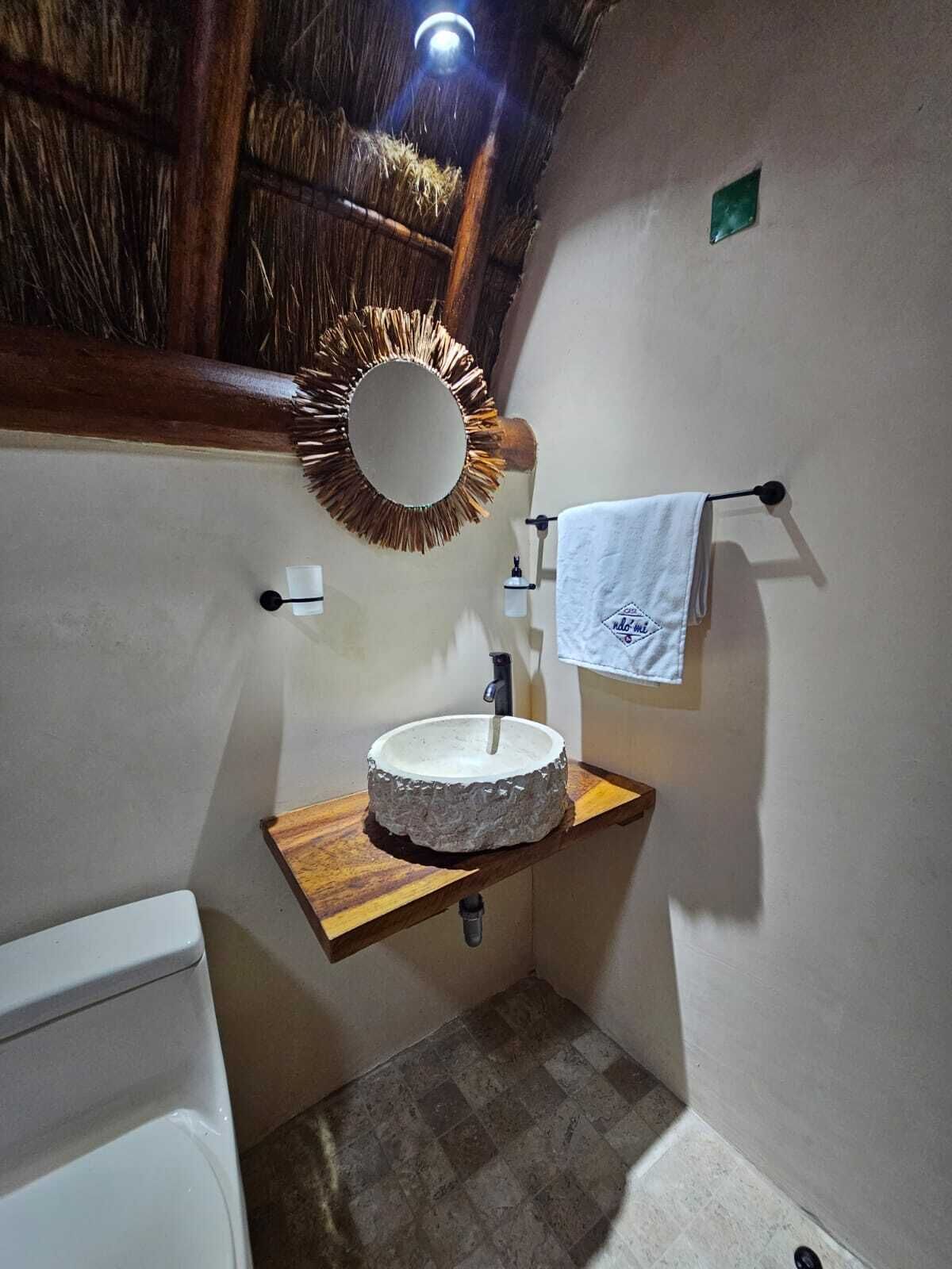 Une salle de bain avec un miroir rond, un lavabo blanc, une étagère en bois, un porte-serviettes et des toilettes.