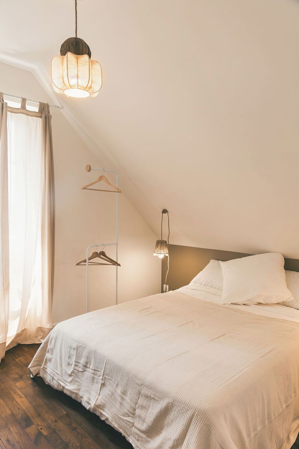 Ein Schlafzimmer mit einem Bett, einer Pendelleuchte, einem Kleiderhaken und einem Fenster mit Vorhängen.
