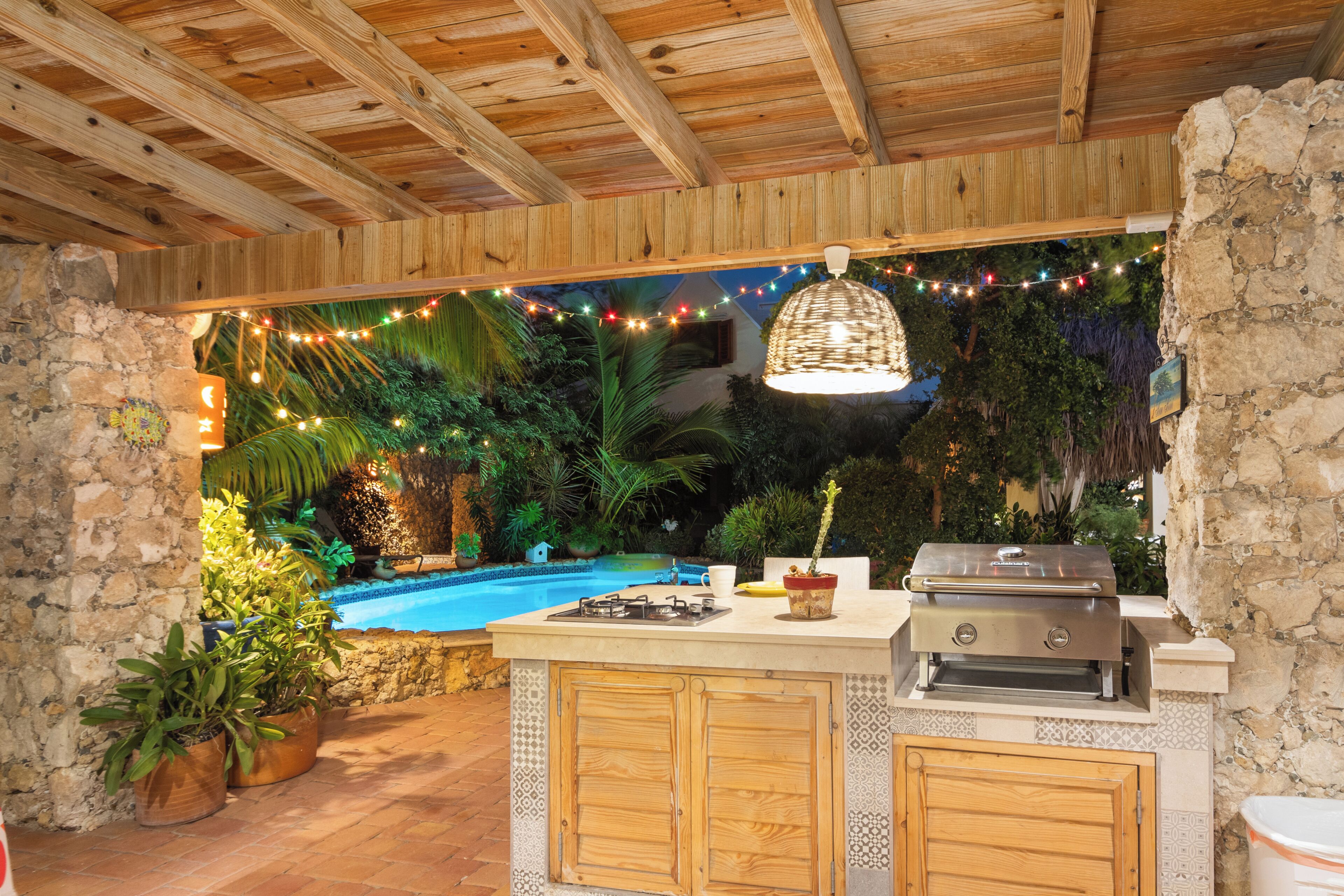 Eine Außenküche mit Grill, Blumentöpfen und einem Pool im Hintergrund.