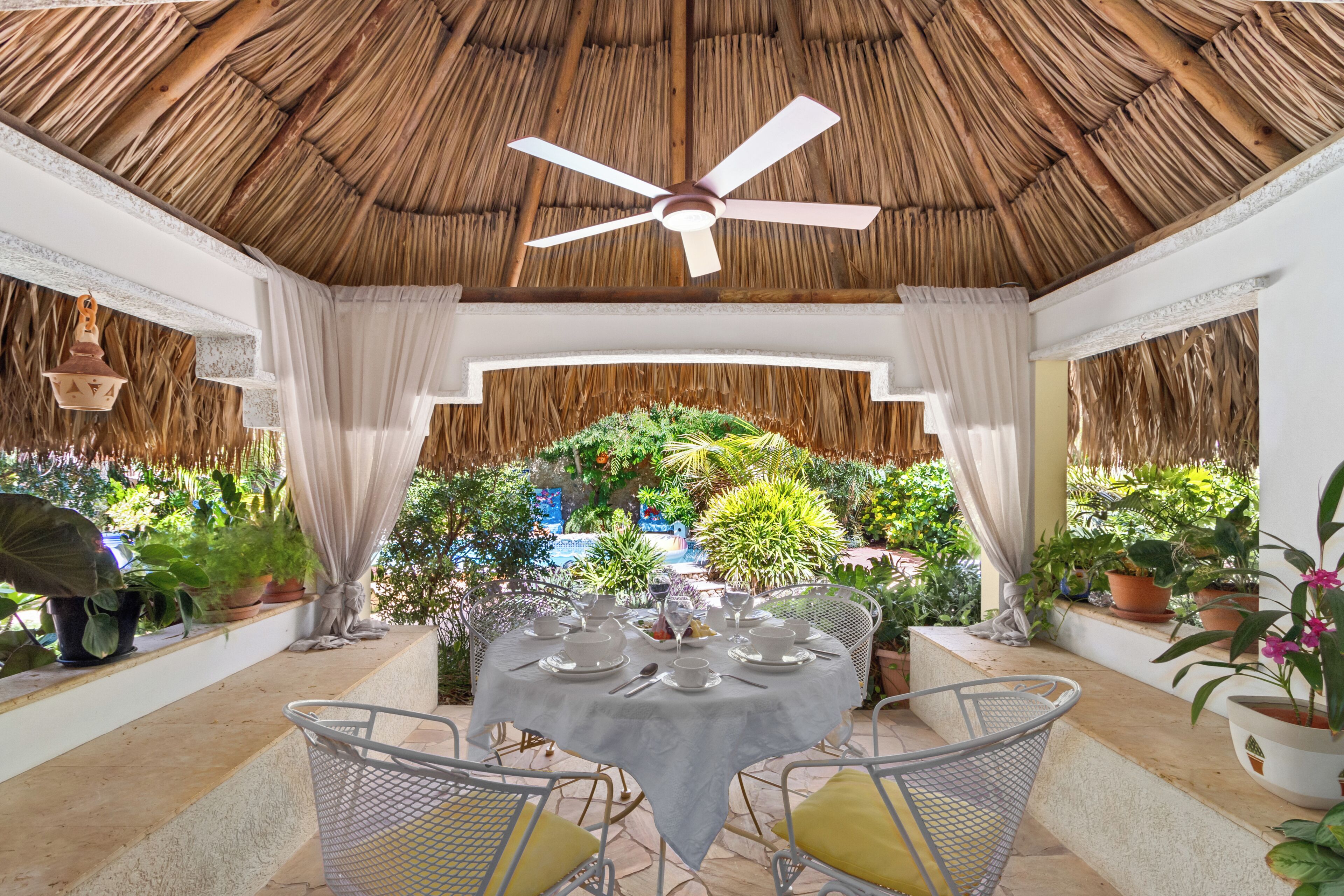 A thatched-roof outdoor dining area with a table set for two, surrounded by potted plants and a view of a lush garden.