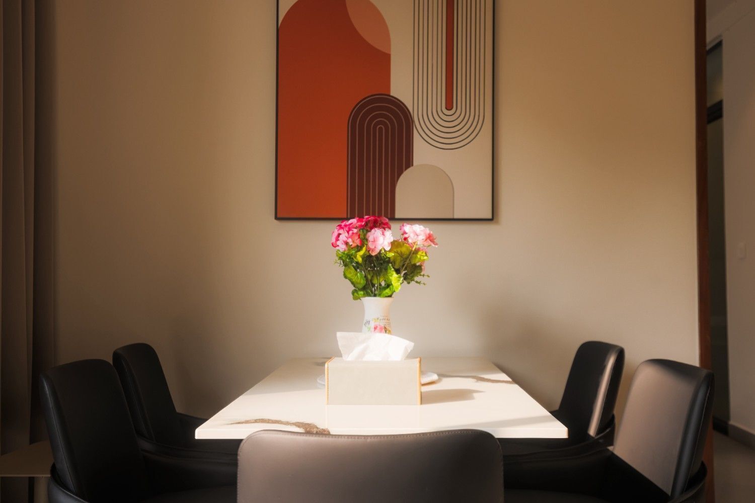 A dining area with a table, chairs, a vase of flowers, and a framed abstract artwork on the wall.