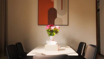 A dining area with a table, chairs, a vase of flowers, and a framed abstract artwork on the wall.