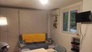 Una habitación con sofá, una mesita, televisión instalada en la pared y ventana con vista a follaje.