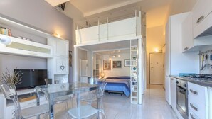 A modern apartment with a loft bedroom, a dining area with clear chairs, a kitchen with white cabinets, and a staircase to the upper level. - Cozy 39m2 studio, ideal for an adult and a child up to 10 years old, located on the ground floor in lively Bolognina district. The location is strategic: the Fair, Tecnopolo & ring road are easily reachable, as are Central Station, the new Municipality h (Bologna)
