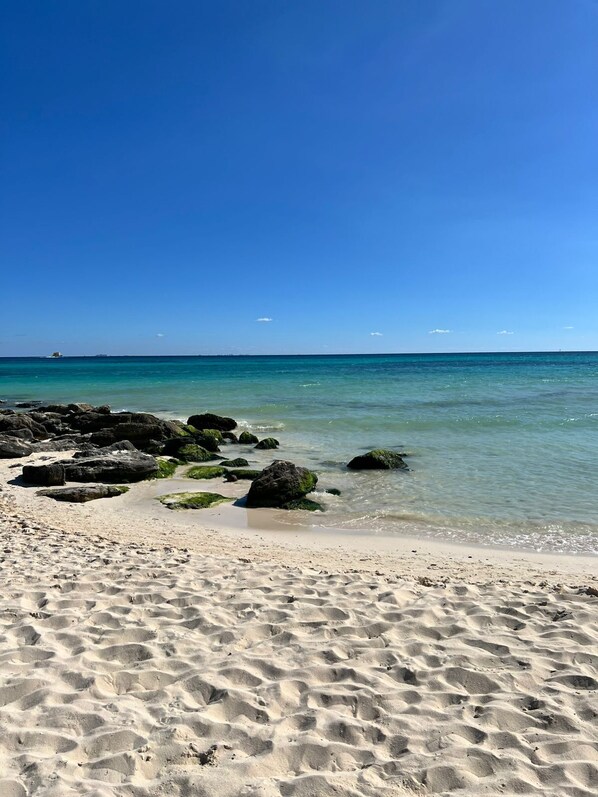 Ein Strand mit klarem Wasser, Felsen und einem Schiffen im Hintergrund.
