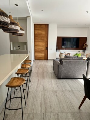 Eine moderne Küche mit Holztür, einer Bar inklusive Barhocker, einem grauen Sofa und einem flachbildfernseher an der Wand.
