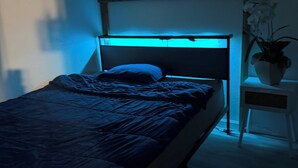 Un dormitorio con una cama, una mesita de noche, una planta en maceta y una ventana con iluminación azul.